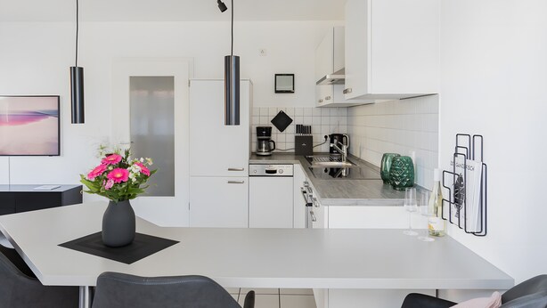 Una cocina moderna con armarios blancos, una mesa de comedor con sillas, un jarrón de flores y una estantería para vinos.
