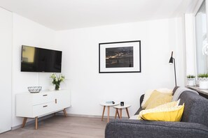 Un salon moderne avec un téléviseur à écran plat, une photo encadrée, un meuble blanc, un canapé gris et une petite table ronde sur laquelle se trouve une télécommande.