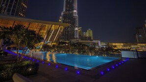 Vista nocturna de un edificio moderno con una aguja distintiva, rodeado de un área de piscina con sillones de descanso e iluminación ambiental azul.