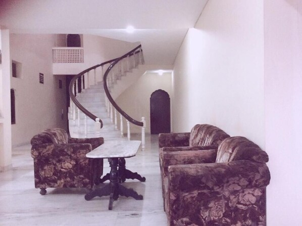 A spacious living area with a marble floor, a central table, and two sofas. A staircase with a wooden railing leads to an upper level. - Hotel Marble Palace (Khajuraho)