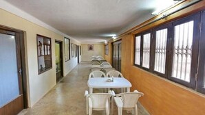 A corridor with a row of white tables and chairs, wooden paneling on the walls, and windows with metal bars. - Hotel Marble Palace  (Khajuraho)
