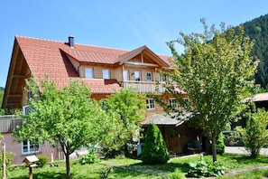 A two-story wooden house with a red-tiled roof, a balcony, and a garden with trees and a small shed. - Apartment 'Kinzigstuben' with Mountain View, Shared Terrace and Wi-Fi (Alpirsbach)