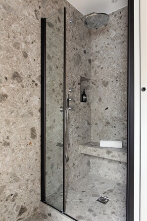 A glass-enclosed shower with a stone wall, a handheld showerhead, and a small shelf with a bottle and a towel. - HIGHSTAY - Appartements de luxe - Arc de Triomphe (Paris)