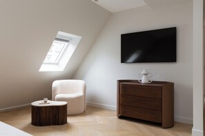 A modern room with a sloped ceiling, a large flat-screen TV mounted on the wall, a round wooden coffee table, a cushioned chair, and a wooden dresser with drawers. - HIGHSTAY - Appartements de luxe - Arc de Triomphe (Paris)