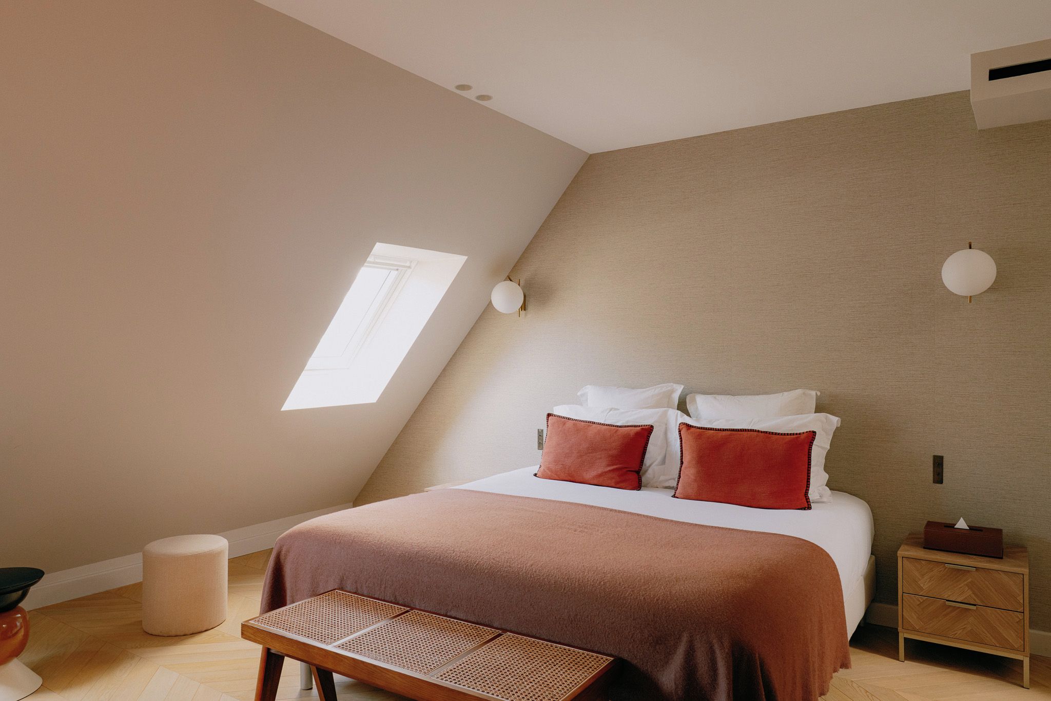 Una camera da letto con soffitto spiovente, un lucernario, un letto con cuscini bianchi e rossi, un comodino con una lampada e una panca in legno.