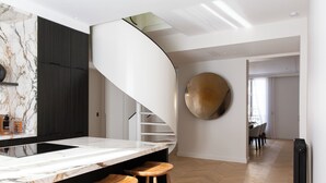 A modern kitchen with a marble countertop, wooden stools, a black vertical panel, and a spiral staircase leading to a dining area. - HIGHSTAY - Appartements de luxe - Arc de Triomphe (Paris)