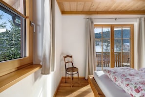 A room with a wooden ceiling, a bed with floral bedding, a wooden chair, and a window with curtains.