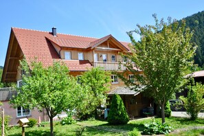 A two-story wooden house with a red-tiled roof, a balcony, and a garden with trees and a small shed.
