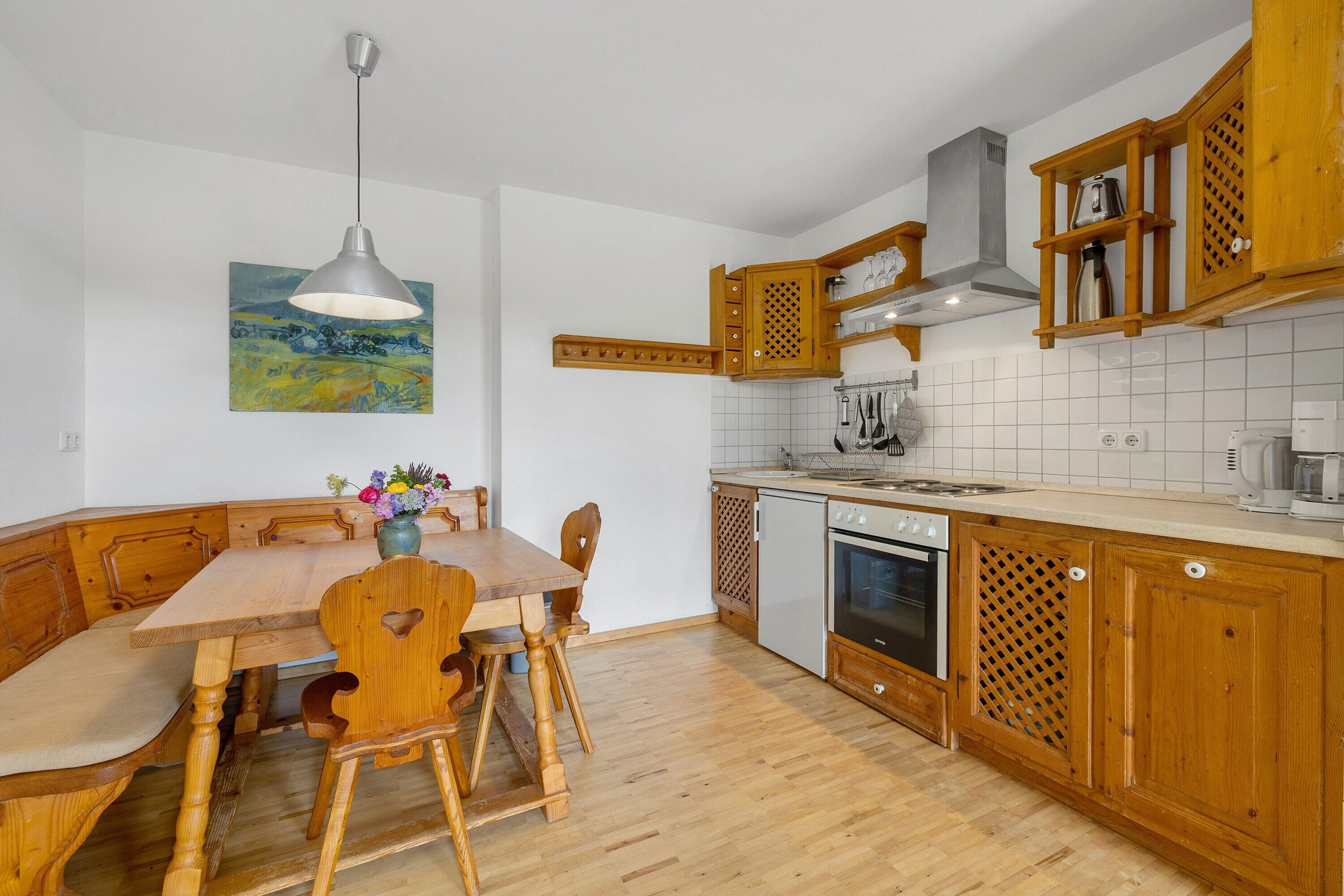 Una cocina con armarios de madera, una mesa de comedor, sillas, una lámpara colgante y un cuadro en la pared.