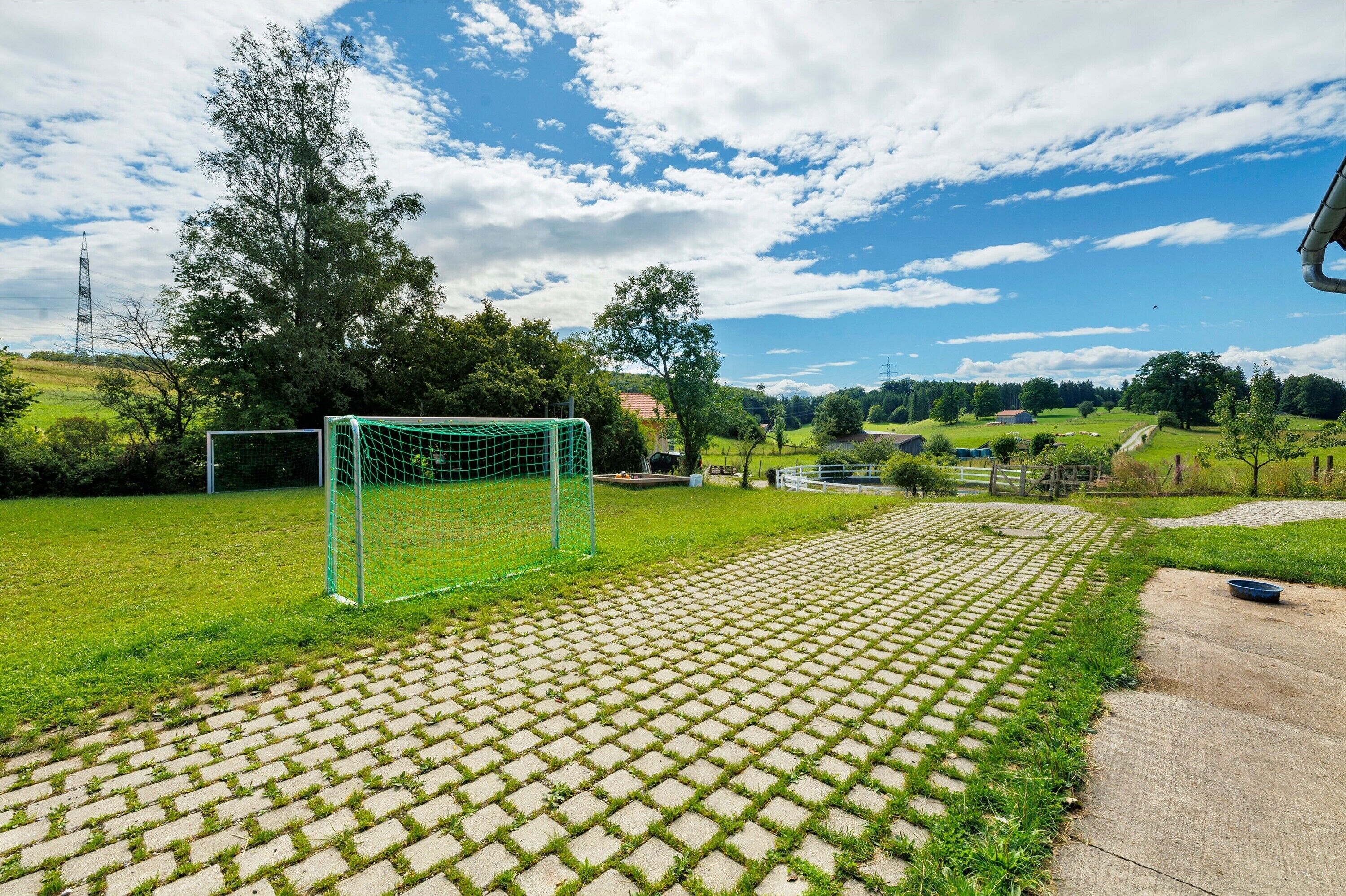A soccer field with a goalpost, a paved walkway, and a grassy area with trees and a clear sky.