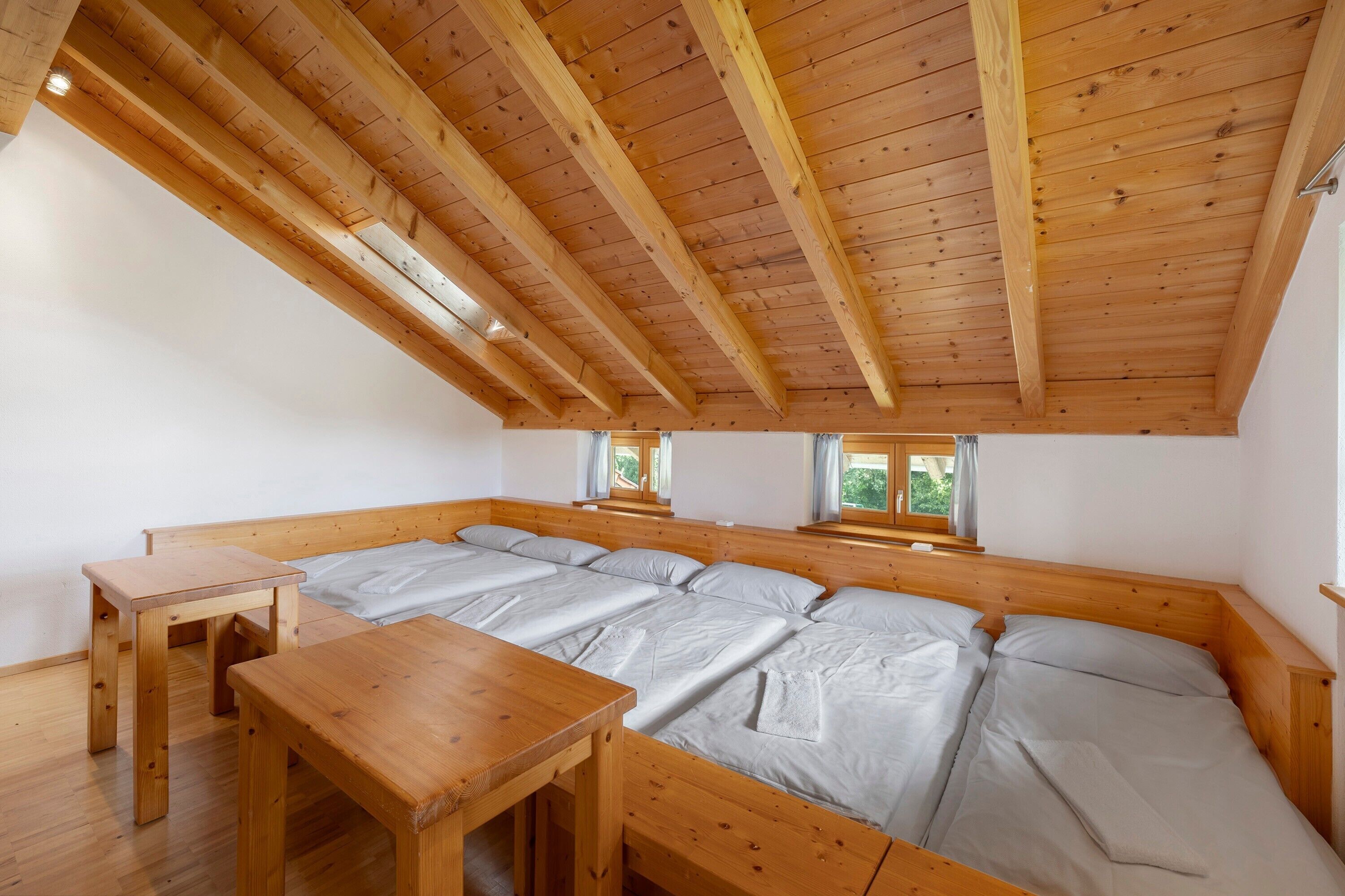 A room with a high wooden ceiling, a large bed with a wooden headboard, a wooden table, and two windows.