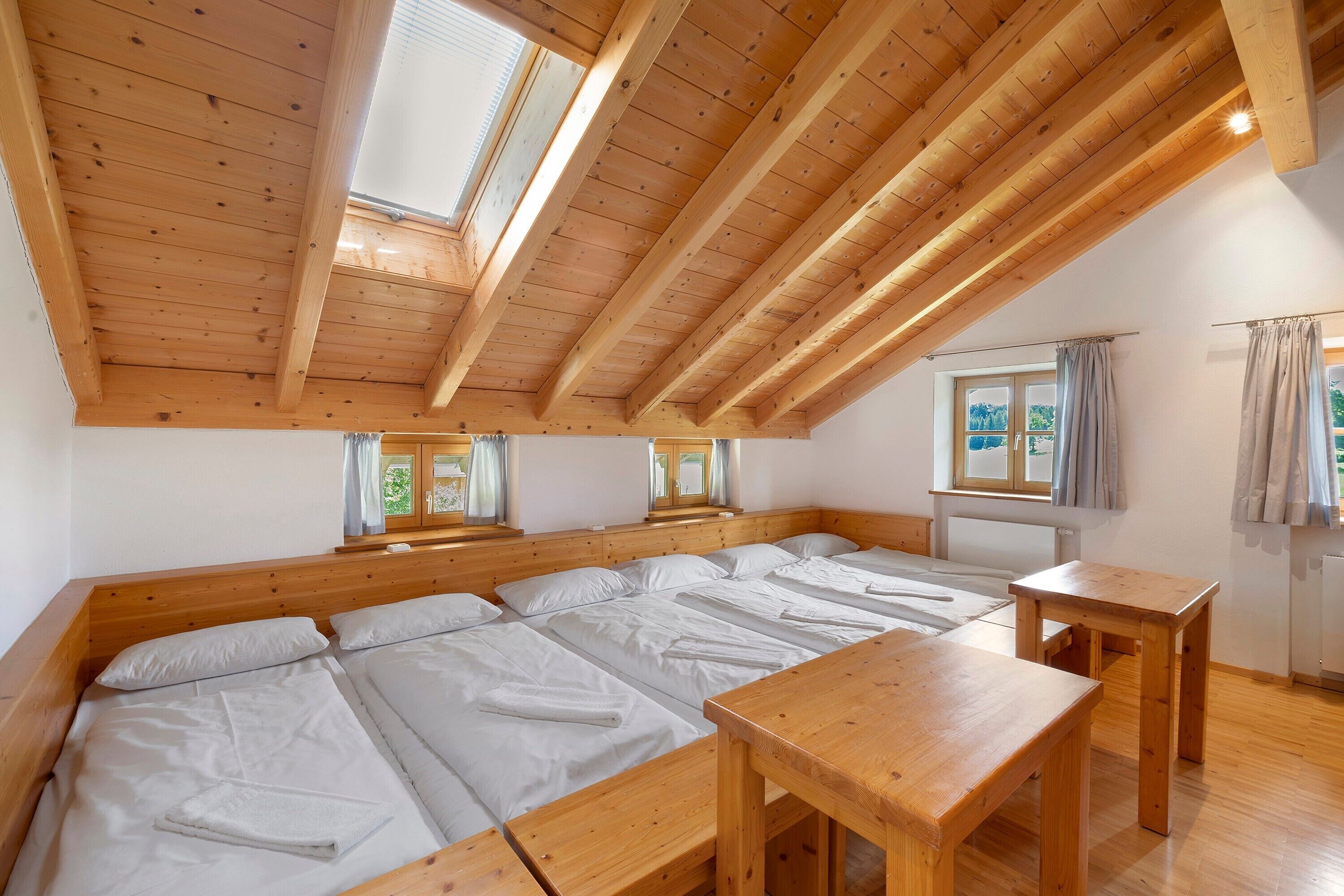 A room with a sloped ceiling, wooden beams, a large bed with white linens, a wooden table, and a window with curtains.