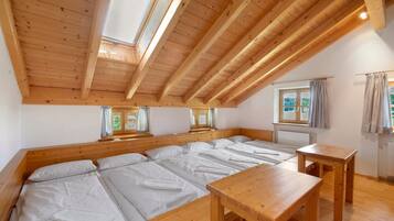 A room with a sloped ceiling, wooden beams, a large bed with white linens, a wooden table, and a window with curtains.