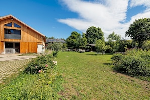 A wooden building with a balcony, a paved area, and a grassy yard with trees and bushes. - Mattress Dormitory 'Yellow' with Shared Terrace & Garden, WITHOUT Kitchen (Seeshaupt)