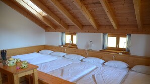 Habitació amb un sostre de fusta alt, un llit gran amb molts coixins, una taula de fusta amb una tetera i dues finestres amb cortines.