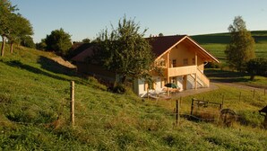 Una casa de fusta amb teulada vermella, envoltada d'un camp herbós i arbres.