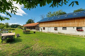 A large barn with a sloped roof, a grassy yard, and a dining area with tables and chairs. - Mattress Dormitory 'Yellow' with Shared Terrace & Garden, WITHOUT Kitchen (Seeshaupt)