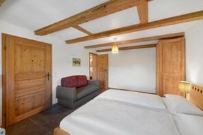 Un dormitorio con cama, vigas de madera en el techo, un sofá con manta roja y negra, y una puerta de madera.