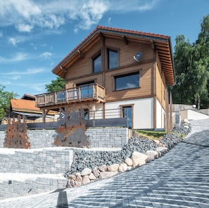 Una casa moderna de dos pisos de madera, con balcón, pared de piedra y camino de entrada pavimentado.