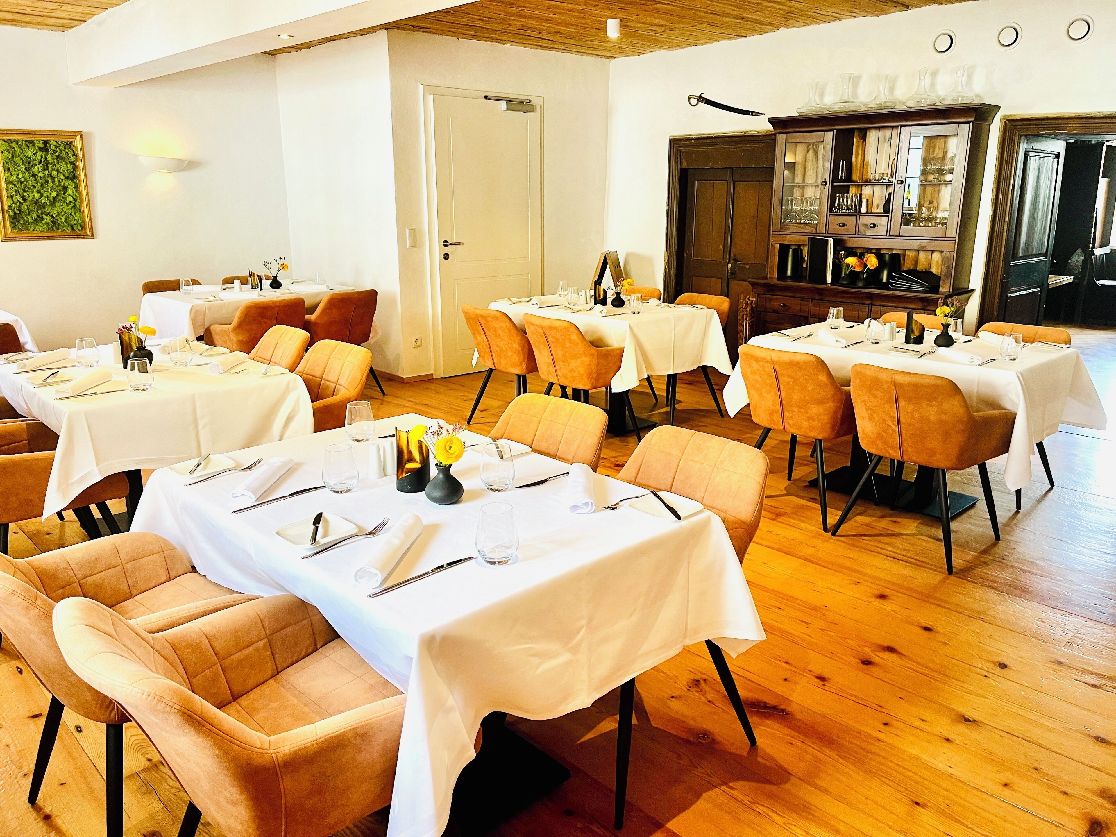A dining area with tables set for four, each with a vase of yellow flowers, in a room with wooden flooring and white walls.