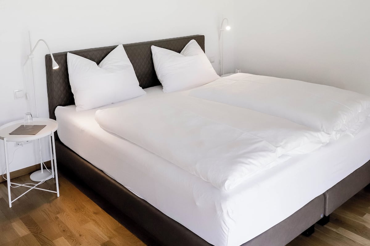 Una cama bien tendida con ropa blanca, un cabecero de madera y una mesita de noche con una lámpara.