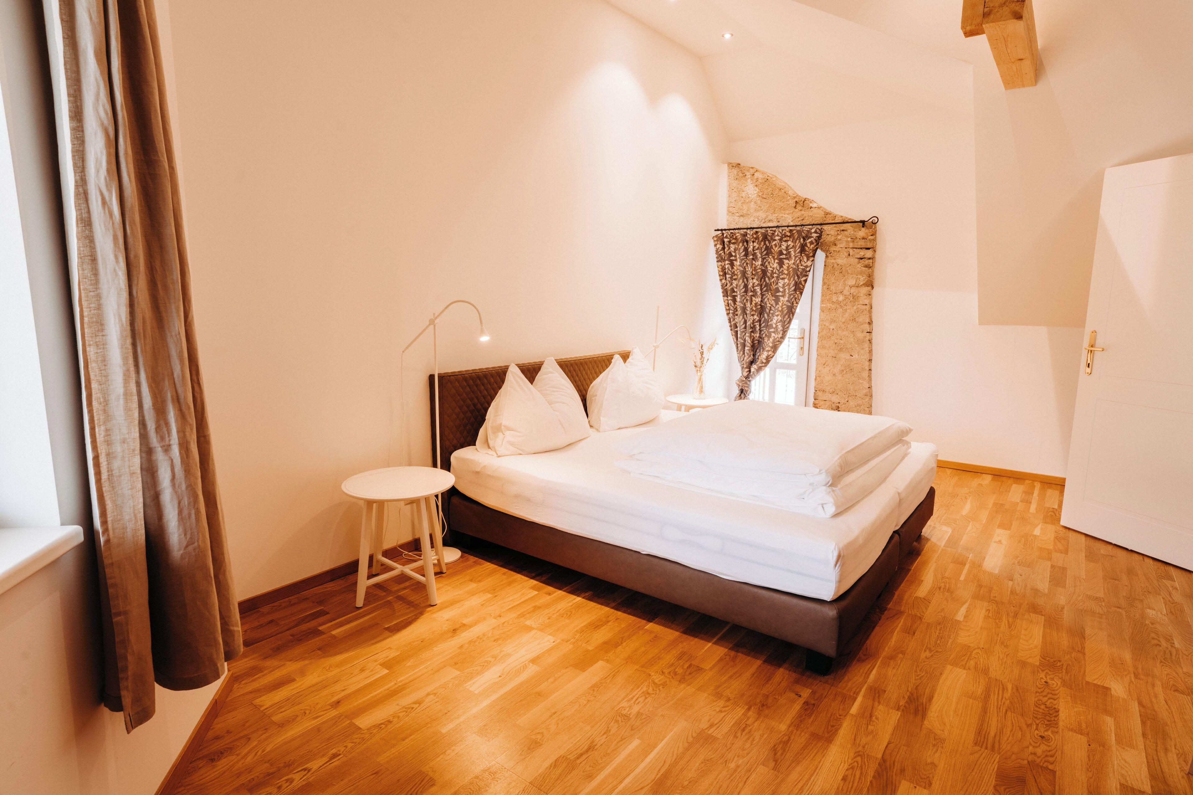 Un dormitorio con cama, una mesita de noche, una ventana con cortinas y suelo de madera.