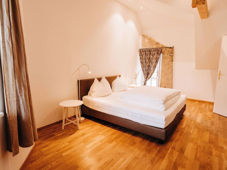 Ein Schlafzimmer mit Bett, kleinem Nachttisch, Fenster mit Vorhängen und Holzboden.