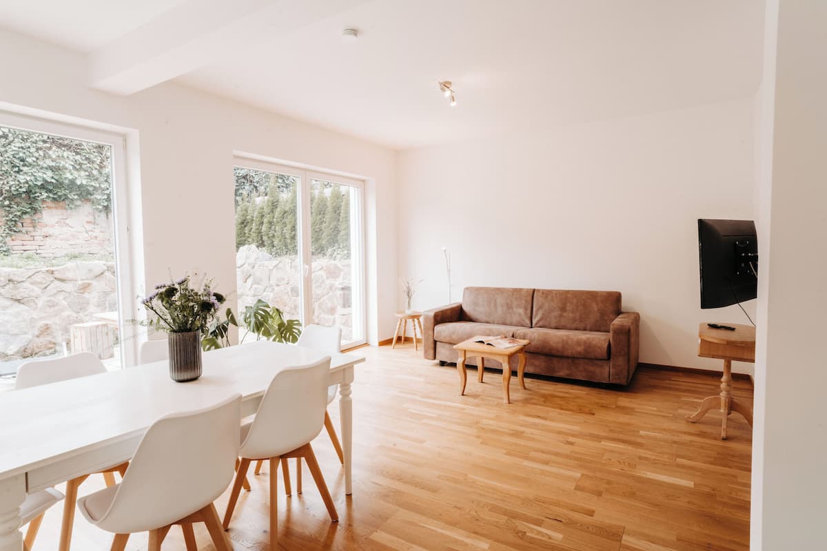 Una sala de estar moderna con piso de madera, una mesa de comedor blanca con sillas, un sofá marrón y un televisor sobre un mueble de madera.