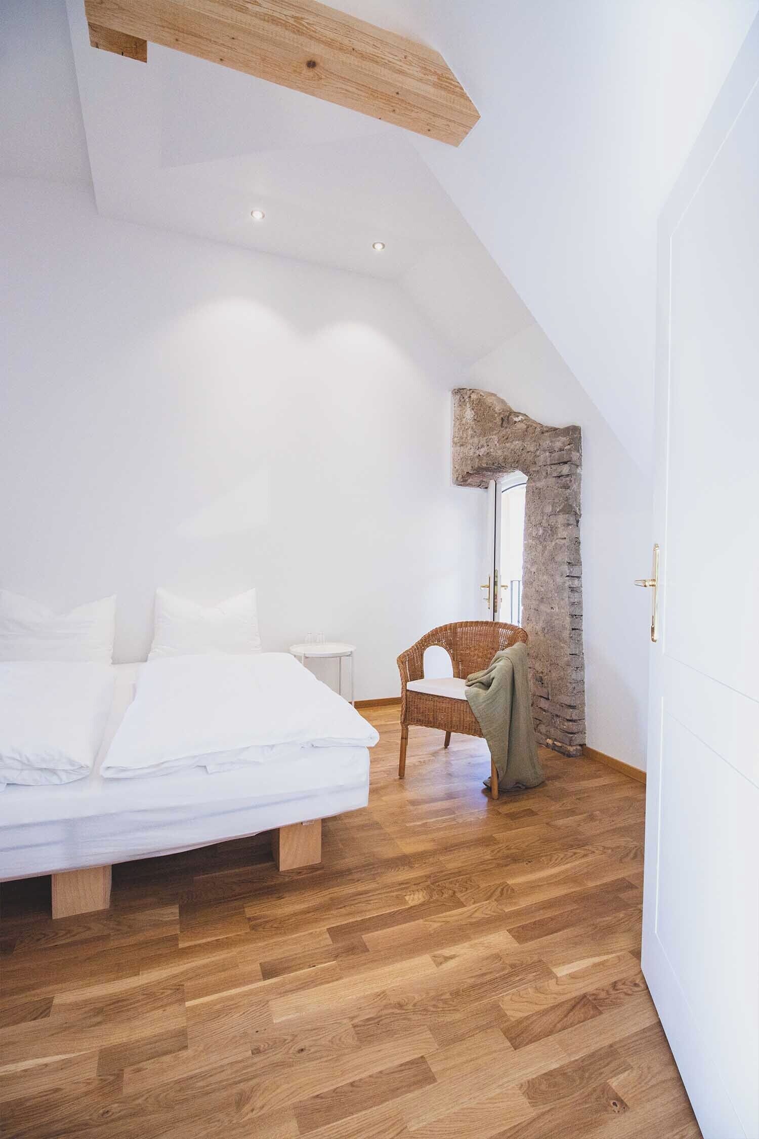 A bedroom with a wooden floor, a bed with white linens, a wicker chair, and a stone wall.
