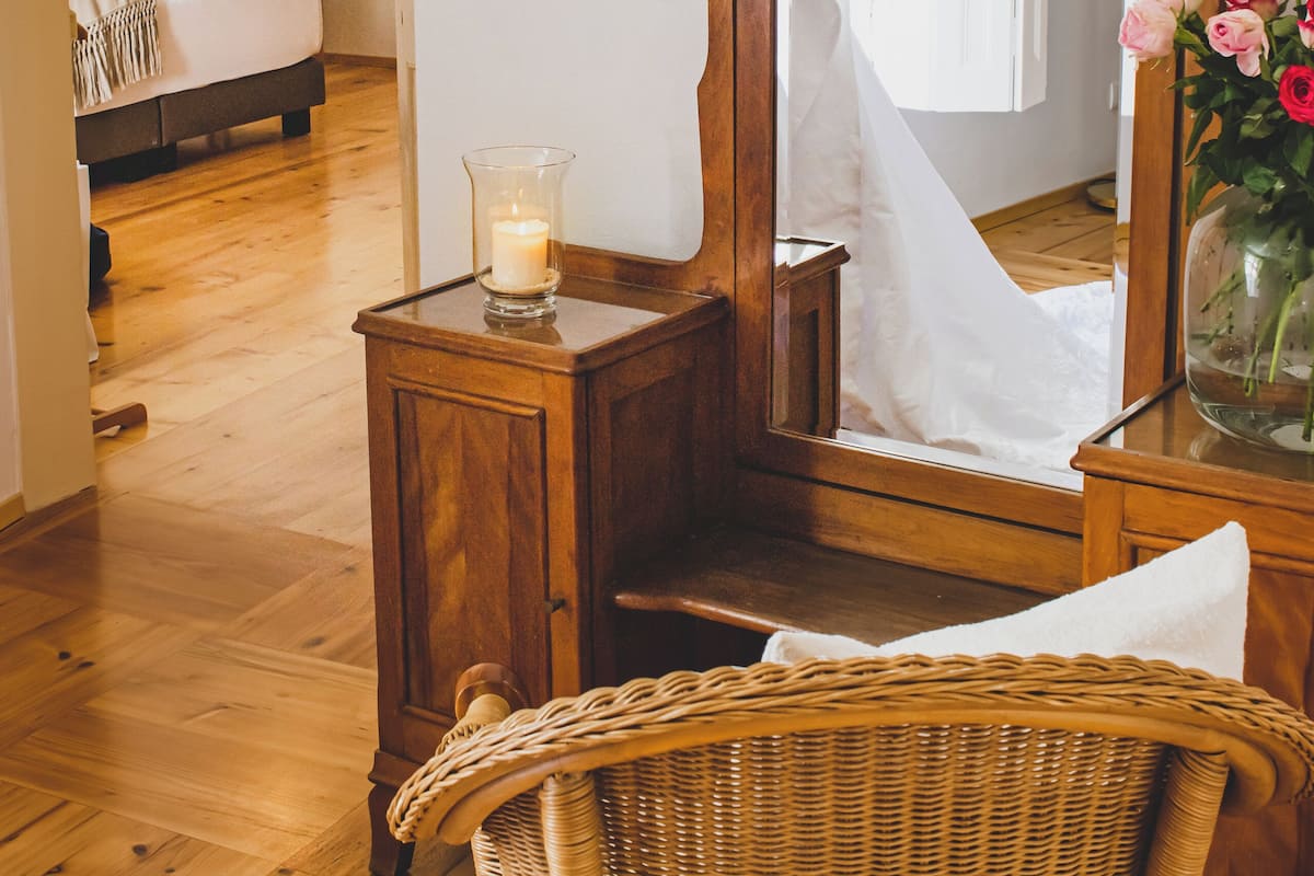 Una habitación con un aparador de madera, una silla de mimbre, un espejo y una mesita de noche con una vela y flores.