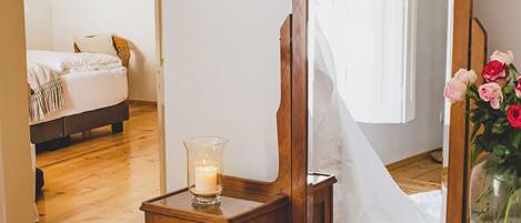Una habitación con un aparador de madera, una silla de mimbre, un espejo y una mesita de noche con una vela y flores.