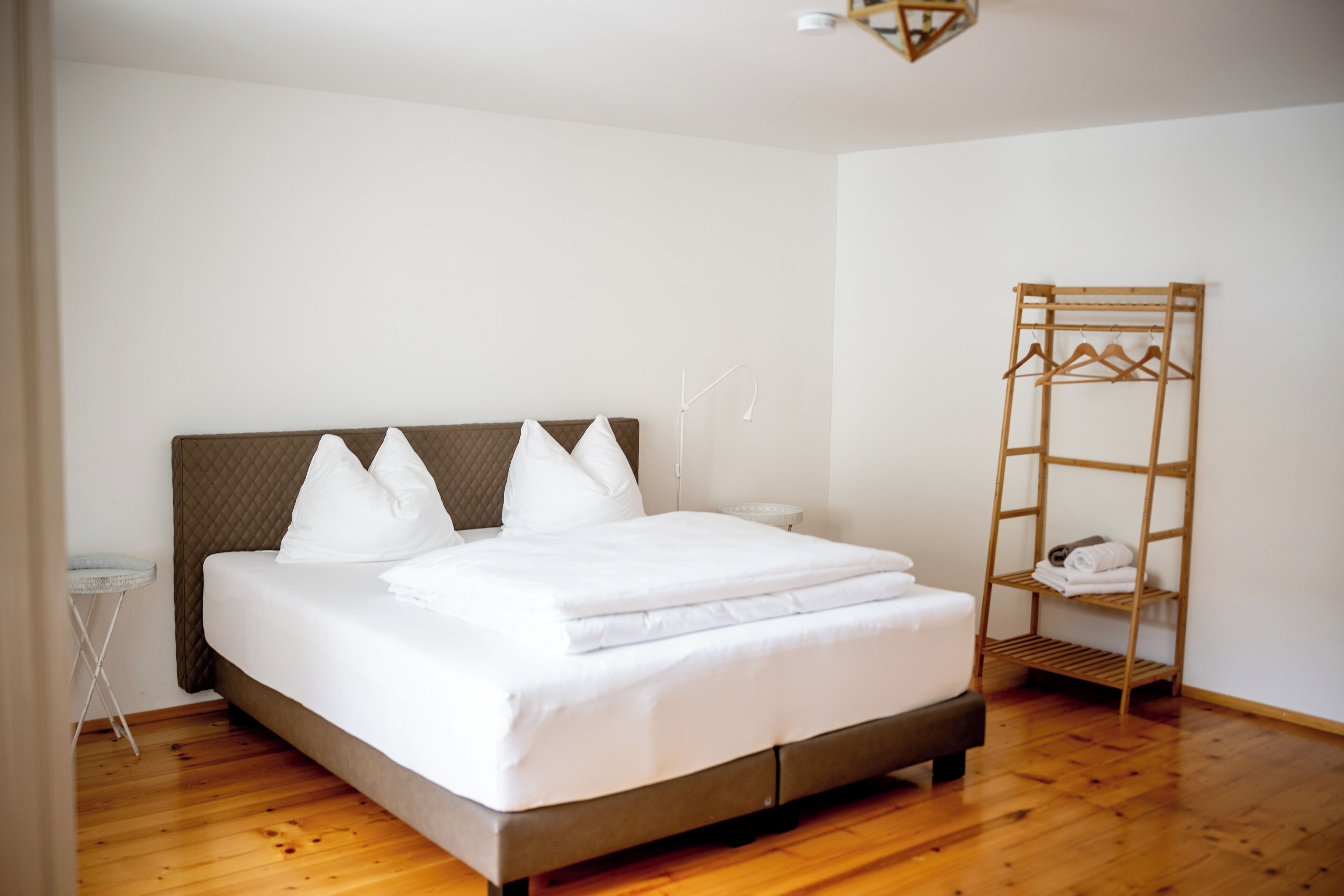 A neatly made bed with white linens and a wooden headboard in a room with wooden flooring and a wooden shelf with towels.
