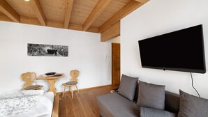 Una habitación con techo de madera, un sofá gris, un televisor montado en la pared y una pequeña mesa de madera con sillas.