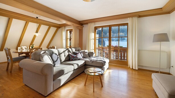 Una sala de estar con un sofá modular, una mesa de centro de vidrio, piso de madera, una zona de comedor con mesa y sillas, y un ventanal con cortinas.
