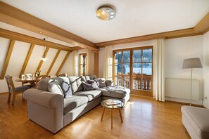 Een woonkamer met een hoekbank, een glazen salontafel, houten vloeren, een eethoek met een tafel en stoelen, en een groot raam met gordijnen.