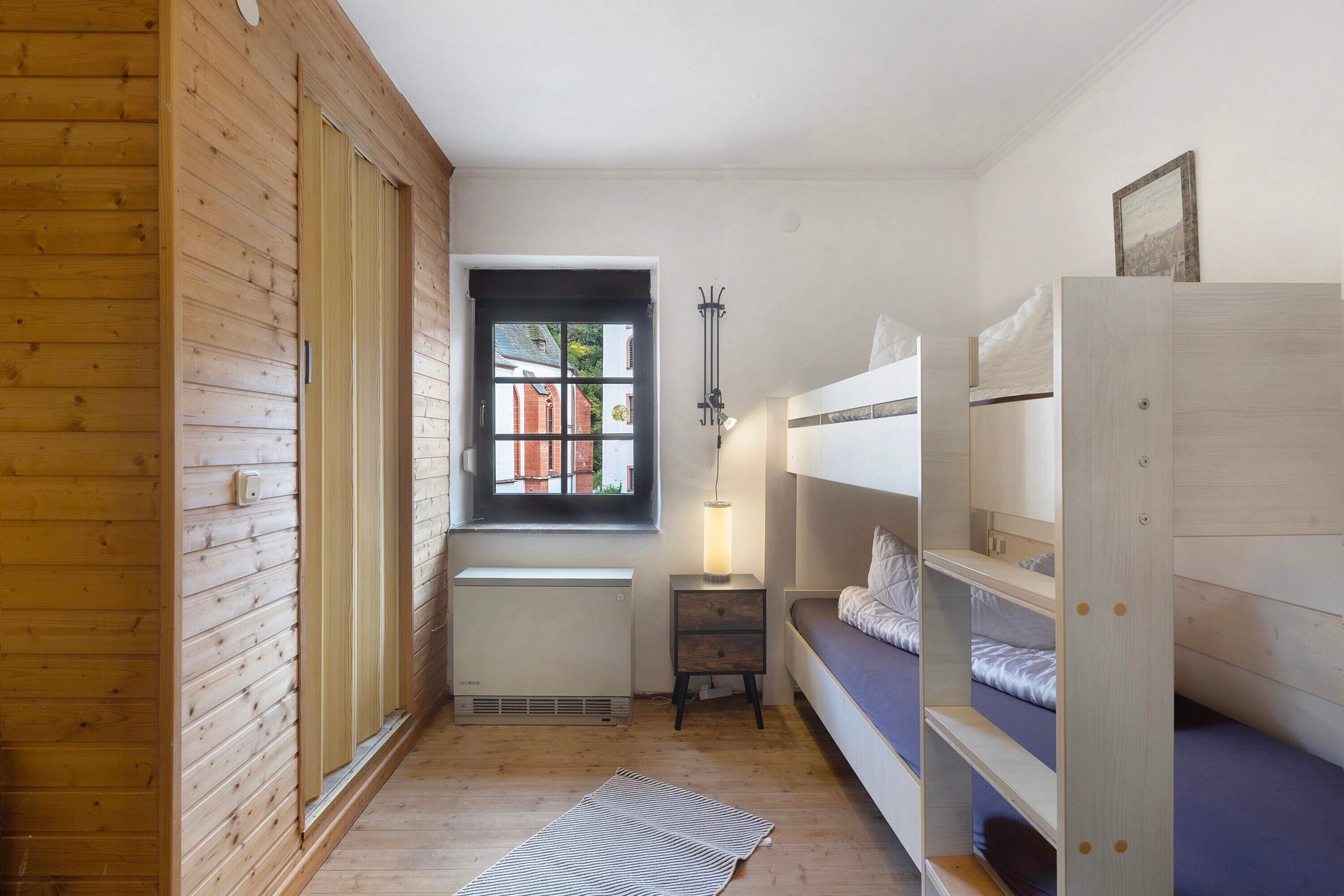Ein Zimmer mit Holzwänden, einem Fenster, einem Heizkörper, einem Nachttisch mit Lampe und einem Etagenbett.