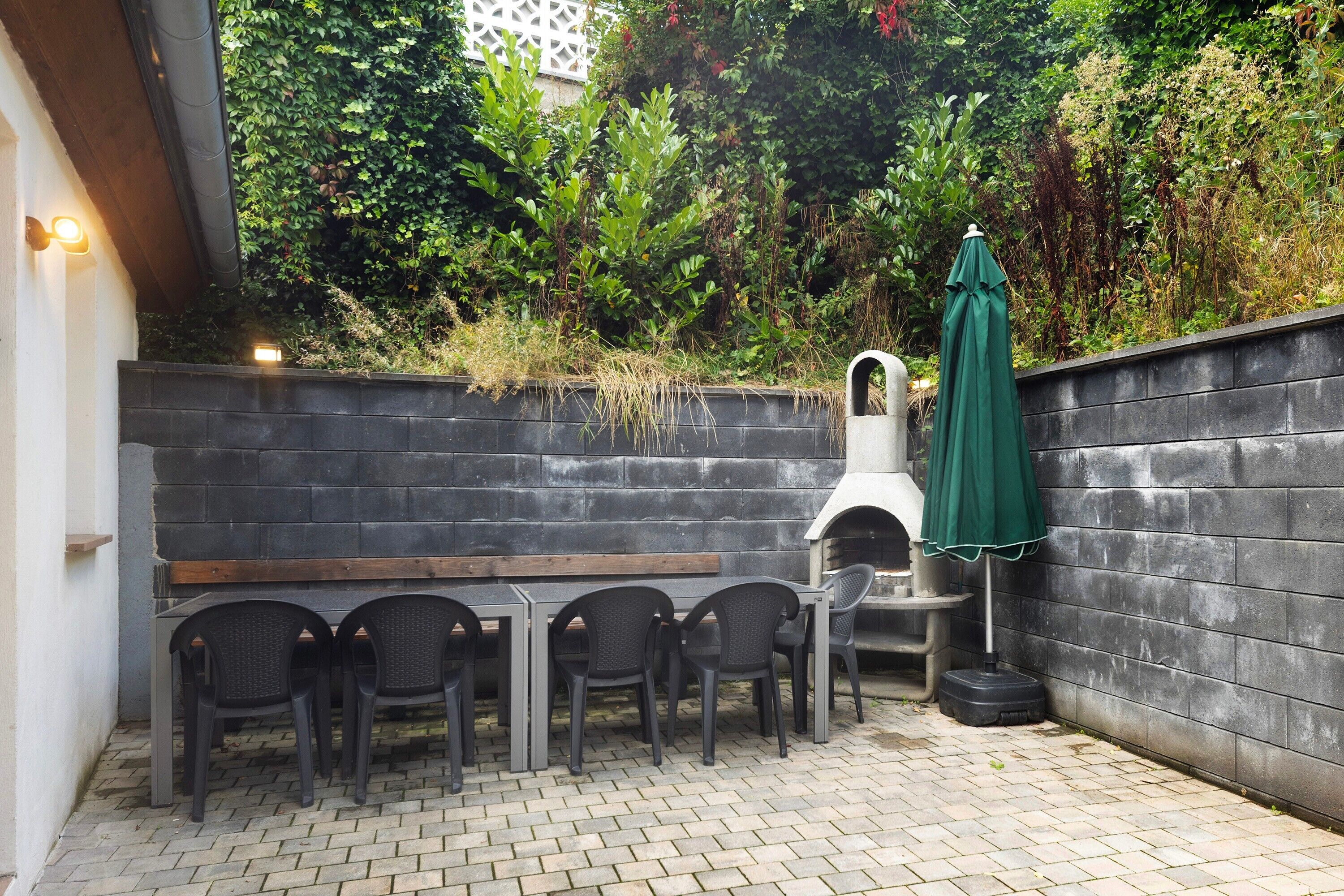 Un patio al aire libre con suelo de ladrillo, una mesa de madera, cuatro sillas, un paraguas verde y un muro de ladrillo.