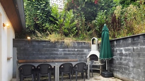 Une terrasse extérieure avec un sol en briques, une table en bois, quatre chaises, un parasol vert et un mur en briques.