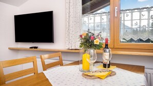 A dining table with chairs, a TV mounted on the wall, a window with a view of mountains, and a vase with flowers. - Holiday Apartment Rubihorn with Mountain View, Balcony and Wi-Fi (Oberstdorf)