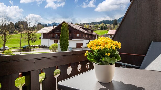 A balcony with a potted yellow flower arrangement, a wooden railing, and a view of a traditional house in the background. - Holiday Apartment Rubihorn with Mountain View, Balcony and Wi-Fi (Oberstdorf)
