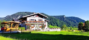 En tvåvånings chalet med en balkong, omgiven av frodiga, gröna fält och berg.