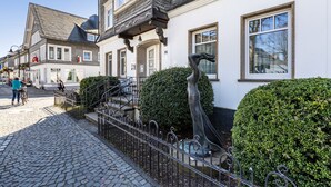 Eine Kopfsteinpflasterstraße mit einem weißen Gebäude, das eine Metallskulptur, eine Treppe und einen Heckenstreifen zeigt.