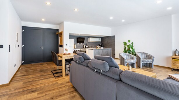 Ein modernes Wohnzimmer mit grauem Sofa, gestreifte Sessel, einem Holzesstisch und einer Küchenzeile im Hintergrund.