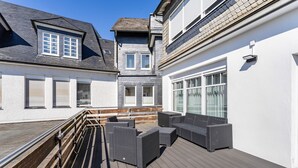 Eine überdachte Terrasse im Freien mit Korbmöbeln, einem Holgitter und Blick auf angrenzende Gebäude.
