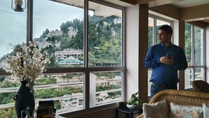 A room with large windows, a vase with flowers, a wicker sofa, and a person holding a phone. - Broadview Homestay  (Darjeeling)