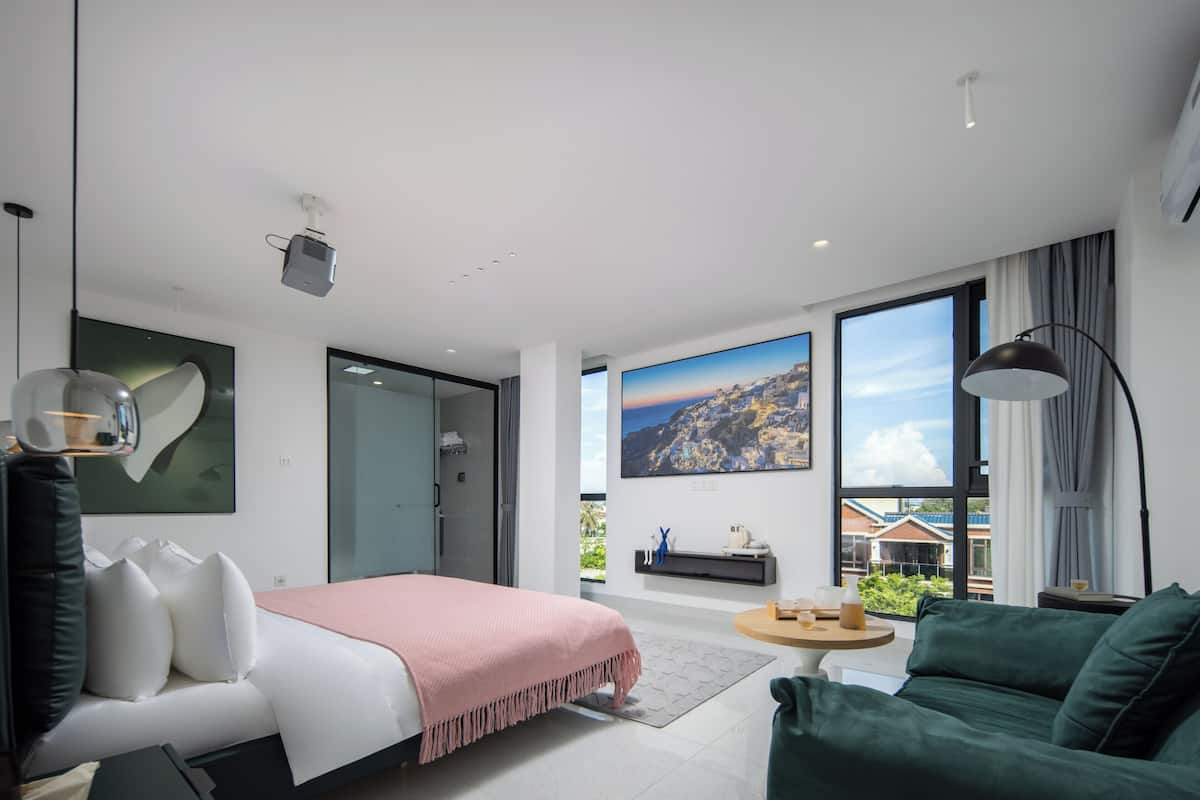 A modern hotel room with a bed, a green armchair, a small table, and a large window with a cityscape view.