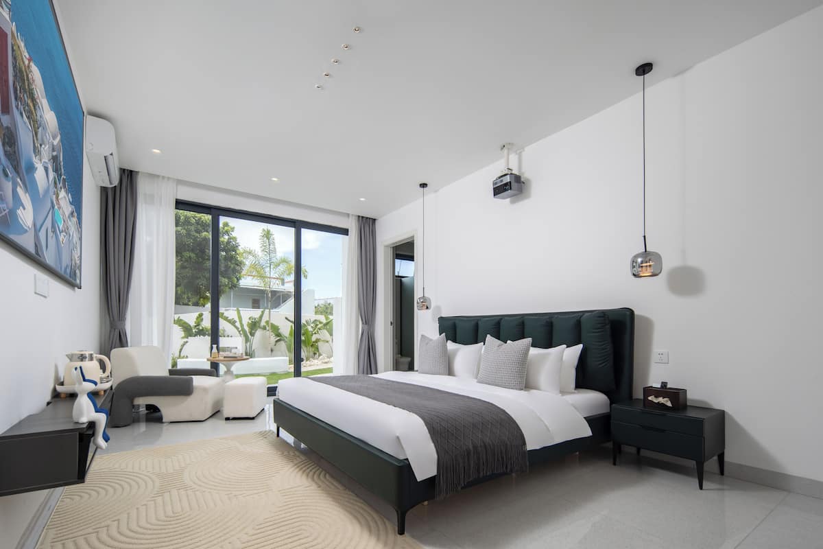 A modern bedroom with a large bed, a seating area with a chair, a desk, and a large window with a view of greenery.