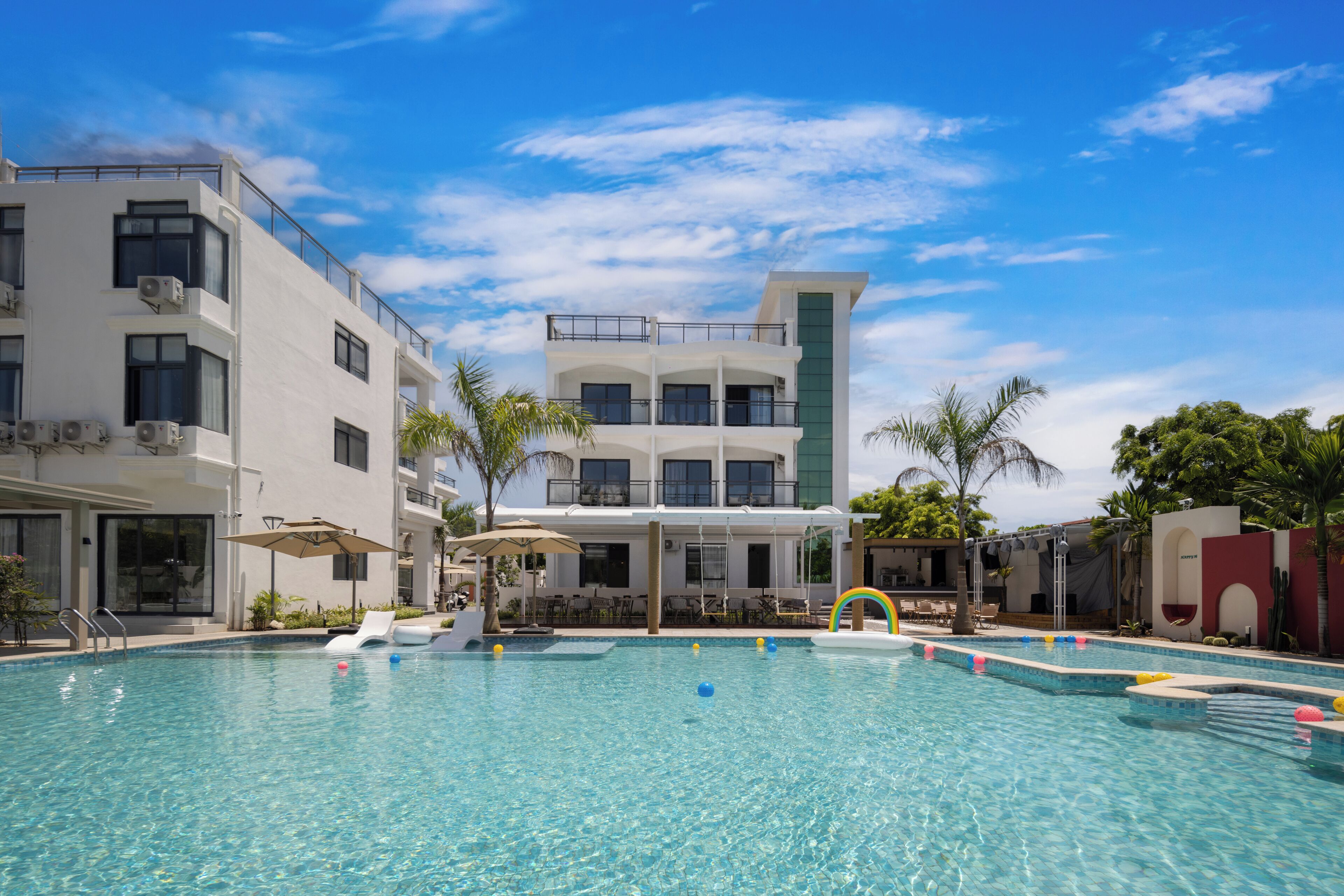 A swimming pool with clear blue water, surrounded by palm trees and a building with balconies.