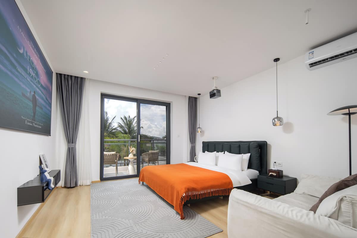A modern bedroom with a large bed, a nightstand, a desk, and a view of the outdoors through a sliding glass door.