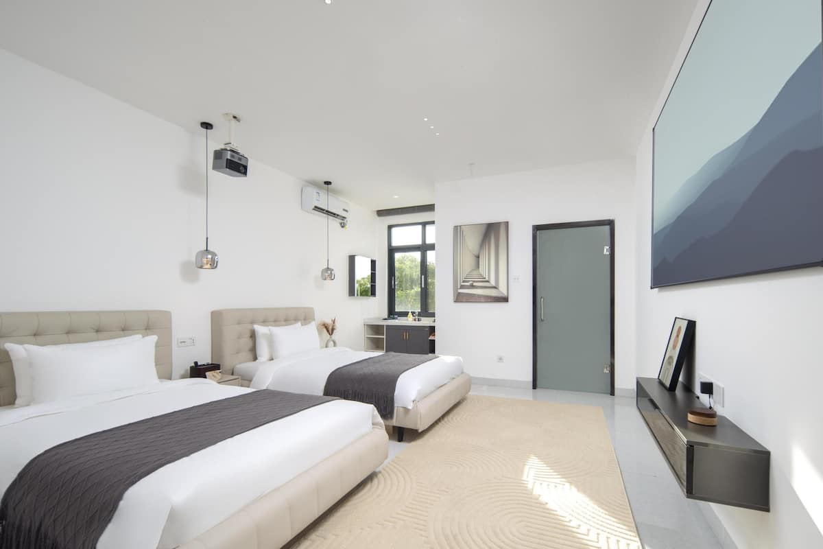 A modern hotel room with two beds, a large window, a flat-screen TV, and a mounted artwork.